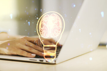 Creative light bulb with human brain hologram and with hands typing on computer keyboard on...