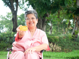 Happy Asian senior woman in pink patient uniform holding credit card and sitting on wheelchair at park. Elderly health care.