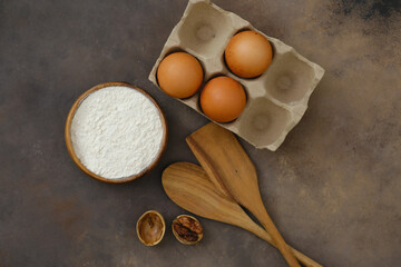 Kitchen utensils and ingredients for baking, spatula, eggs, flour on brown background.