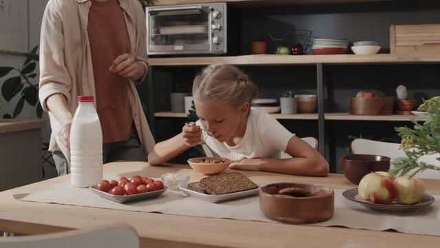 Preteen girl sitting at table in kitchen starting her breakfast while her mother going to cook something else