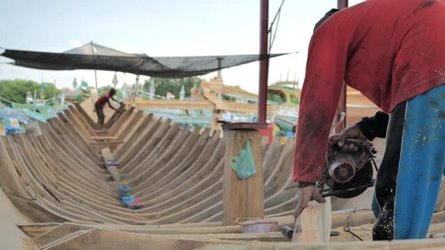 A shipbuilder cuts the timber of a traditional ship's frame on the coast of Indonesia with other workers working on the deck of a ship in a crowded harbor area