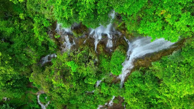 The drone's lens pierces the dense, emerald canopy, revealing a clandestine waterfall tucked away in the tropical rainforest. A surreal, hidden wonder. Thailand.
