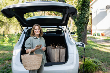 Woman with shopping bag next to a charging electric car in the yard of a country house