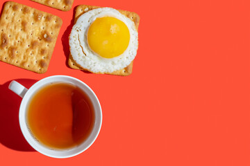 Cup of tea, crackers, sunny side up egg on red background with copy space. Food for breakfast.