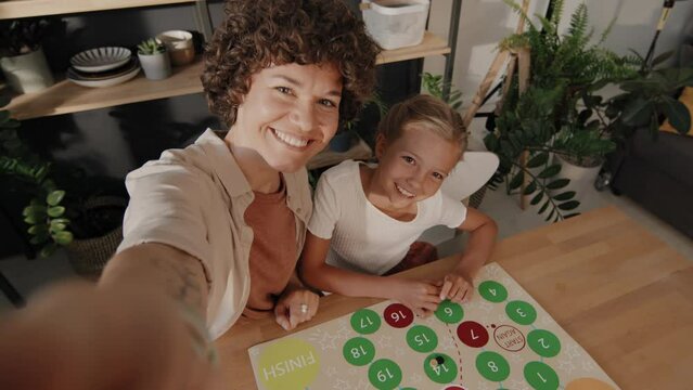 High angle slowmo of cheerful woman holding camera taking selfies with her preteen daughter while playing board game in kitchen