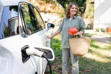 Woman with shopping bag and pumpkins next to a charging electric car in the yard of a country house