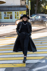 An attractive young woman crosses the road at a pedestrian crossing. A girl in stylish clothes, in a black trench coat and hat