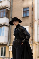Fototapeta na wymiar Portrait of an attractive and young blonde woman in a black hat and a leather trench coat while walking around the city. The concept of style and fashion