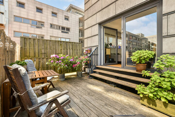 a wooden deck with chairs and potted plants in the foreground, on a sunny day at an apartment building - obrazy, fototapety, plakaty