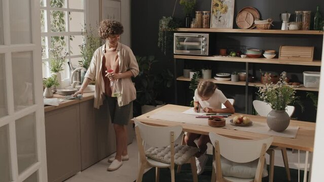 Long shot of woman and daughter spending rime in kitchen, mom doing chores and girl drawing picture