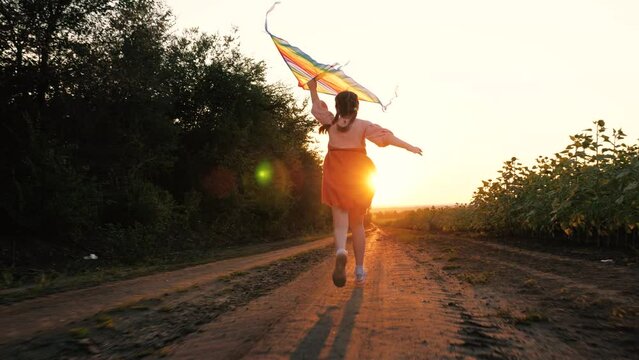 Joyful little girl with kite runs along rural road at back sunset light