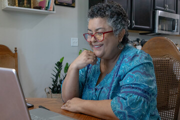 Senior Hispanic woman smiling and talking on video call with family, while sitting at kitchen table - obrazy, fototapety, plakaty