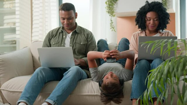 Addicted family use gadgets devices little kid child son boy lying upside down on sofa play video game on mobile phone smiling African American parents working laptops different gen using modern tech