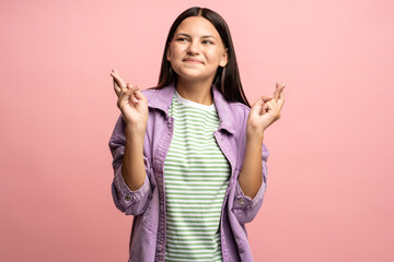 Teenage girl smiling dreaming with fingers crossed. Brunette schoolgirl hopes for good luck, folding hands in happy gesture. Teenager worried, anticipating victory. Isolated on pink studio background.