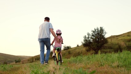 Attentive father teaches preschooler girl to ride bicycle in sunset field