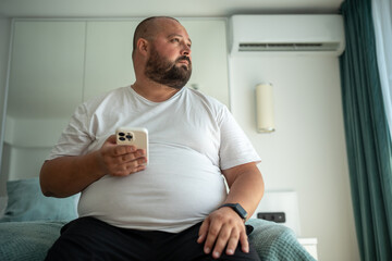 Obese unshaved man with phone and fitness tracker looking at window. Overweight middle-aged man thinks on weight reduction, diet and calories calculation. Weight control and healthy food concept - obrazy, fototapety, plakaty