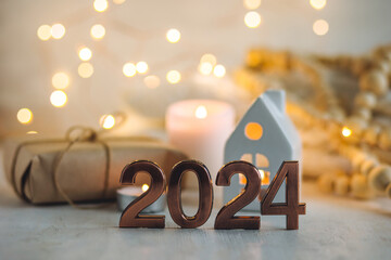 Happy new year 2024 postcard. Golden digits in front of Christmas lights, candles, gift box. White wooden background. Copy space for greeting text