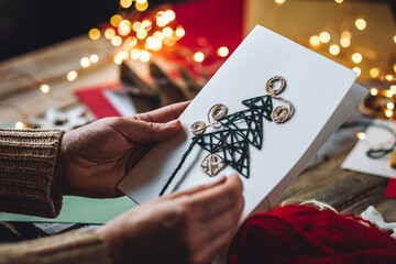 Woman doing handmade Christmas craft card with paper and wool thread. Cozy home atmosphere, lights,...