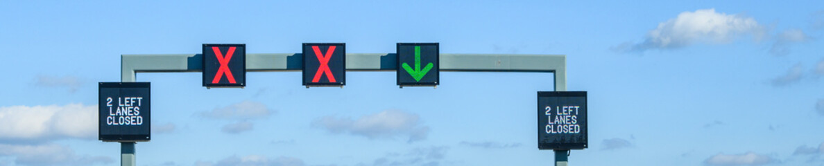 Overhead electronic highway sign, 2 left lanes closed, red x and green arrow, on a sunny blue sky...