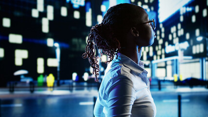 Smiling african american woman wandering around city boulevards during nighttime, enjoying life. Businesswoman strolling around in office buildings urban center, surrounded by bokeh street lights - obrazy, fototapety, plakaty