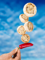 Stuffed mushrooms balancing on a finger and red chilly pepper, blue cloudy sky background. Cooking...