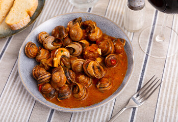 Roasted snails in the tomato sauce on a plate served with cutlery near to pieces of bread on the table in the Spanish cafeteria - Powered by Adobe
