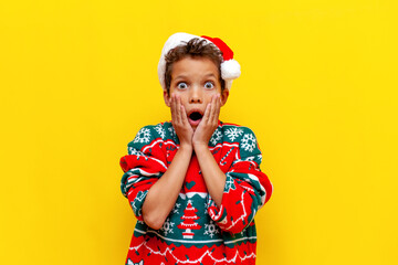 shocked african american boy in christmas clothes wonders with open mouth on yellow isolated...