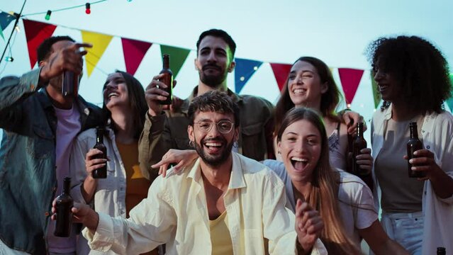 Slow motion of joyful guys and ladies having fun dancing and drinking with raised arms enjoying at the rooftop party. Excited group of young friends holding beer bottles and laughing together. High