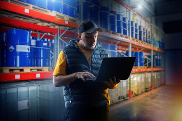 Man with laptop inside logistics warehouse. Storage racks with barrels and boxes. Man works in...