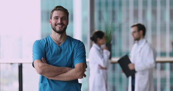 Handsome positive male physician GP wear blue uniform pose in modern clinic office smile feels satisfied with career. Professional medical services, vocation, profession, reliable physician portrait