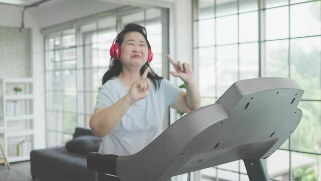 Happy Asian senior woman jogging and listening to music on treadmill at home.