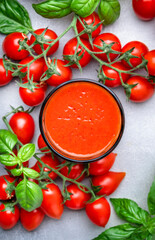 Freshly squeezed tomato juice glass and red cherry tomatoes with green basil leaves on gray table background, top view