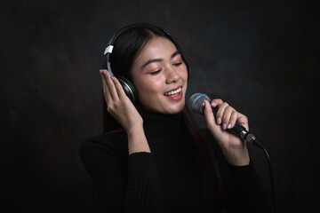 Beautiful young woman with headphones singing in microphone in studio with black wall on the...