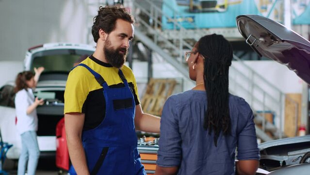 Engineer in auto repair shop showing client malfunctions found inside car after thorough inspection. Repairman telling african american woman vehicle diagnostic after checking for broken parts