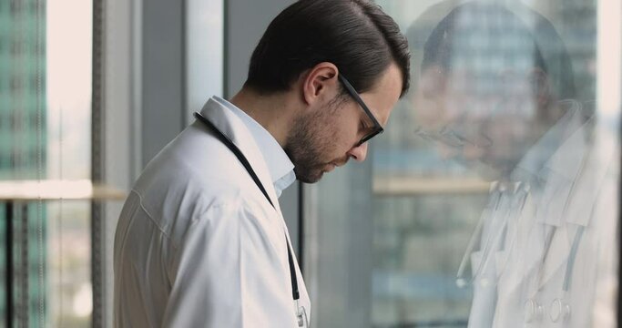 Close up young 35s desperate or fatigued male therapist in white uniform stands near window with bowed head feels concerned, looks terribly upset due to medical error, overloaded. Professional failure