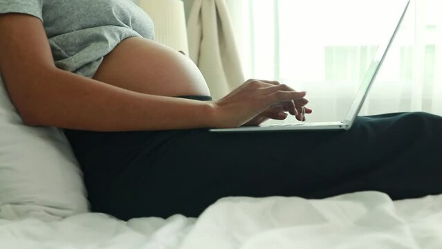 Half Thai African American mother sits on the bed and spends her free time typing on the keyboard on a laptop computer online internet looking for medical knowledge about pregnancy.
