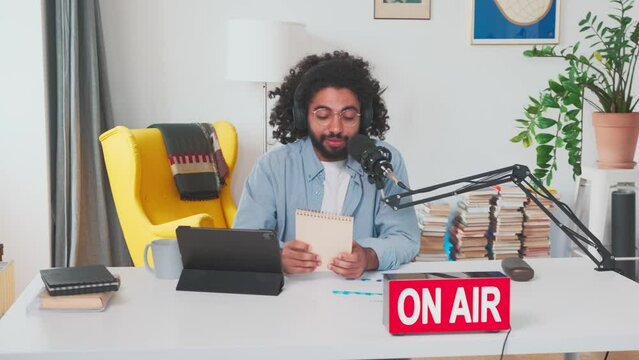 Young charismatic Arabian man blogger records audio podcast for subscribers and holds notepad with broadcast plan and main points of monologue sits in home studio at table with microphone and tablet.