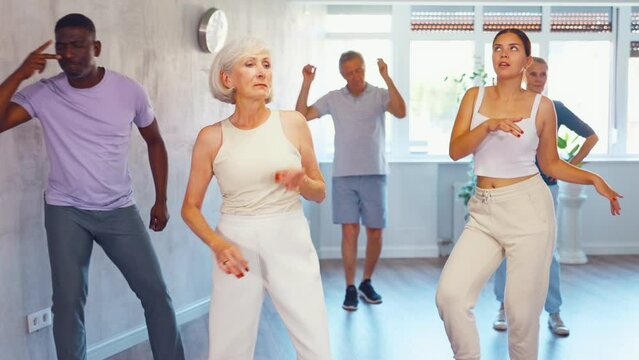 Young girl coach conducts classes for elderly in fitness studio. Female instructor teaches senior students of different ages dance class basic movements of contemporary dances