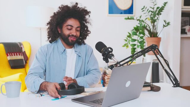 Young happy Arabian man coach takes off headphones and looks at camera after finishing online lecture or recording webinar dressed in casual shirt sits at table with microphone and laptop in office.