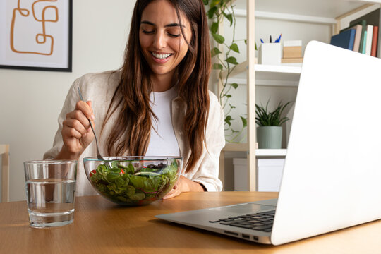 Caucasian woman eating healthy salad for lunch while working with laptop at home office.
