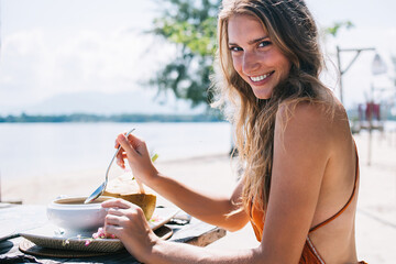 Cheerful adult lady eating while sitting at table on ocean sand beach in fair weather