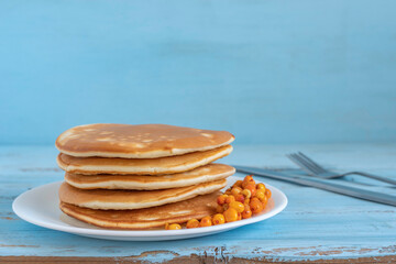 Pancakes and sea buckthorn berries on ceramic plate over blue wooden background.
