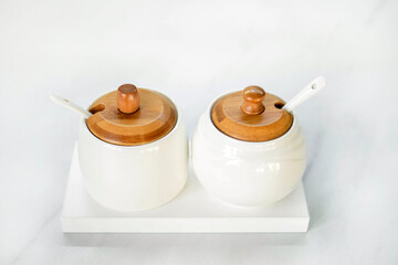 A salt shaker and a sugar bowl in white with wooden lids stand on a white table. Elements of decor in a modern interior. Modern White Tableware