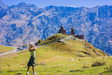 Fototapeta premium A young traveler walks along the path to the Gergeti Temple. A woman enjoys a beautiful view of the Trinity Church in Gergeti. Georgia, Stepantsminda, Gergeti