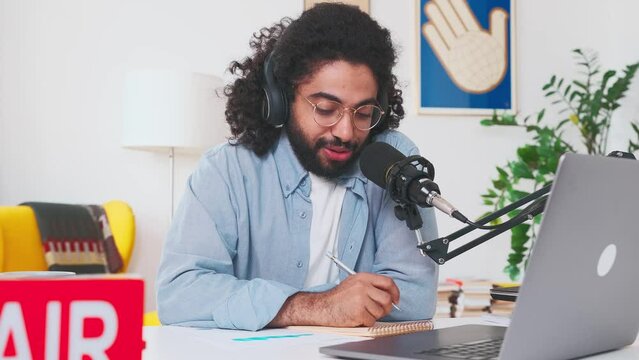 Young curly Arabian man influencer puts on headphones starting to record online podcast or webinar recording with training tips and life hacks for subscribers sits at table in home recording studio.