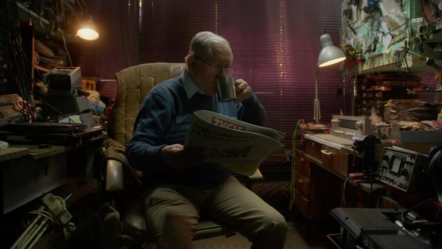 Elderly man reads newspaper and drinks from cup in cluttered room