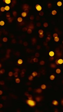 Vertical video. Bokeh light. Blur sparks. Confetti rain. Defocused golden orange glowing glitter circles flying on dark black free space abstract background.