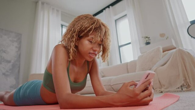 woman dressed bright activewear lying on mat after workout holding smartphone using sport app
