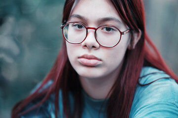 Fototapeta premium Portrait of a sad girl with glasses. A thoughtful red-haired teenage girl in a blue T-shirt looks at the camera..