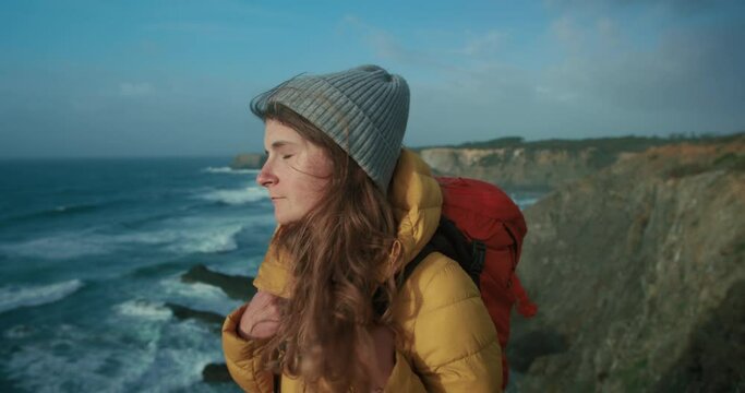Cinematic portrait of young woman turn around and look at camera, modern female hiker in yellow down puffer jacket, red backpack and grey knit beanie. Adventure seeking traveller on hiking trail